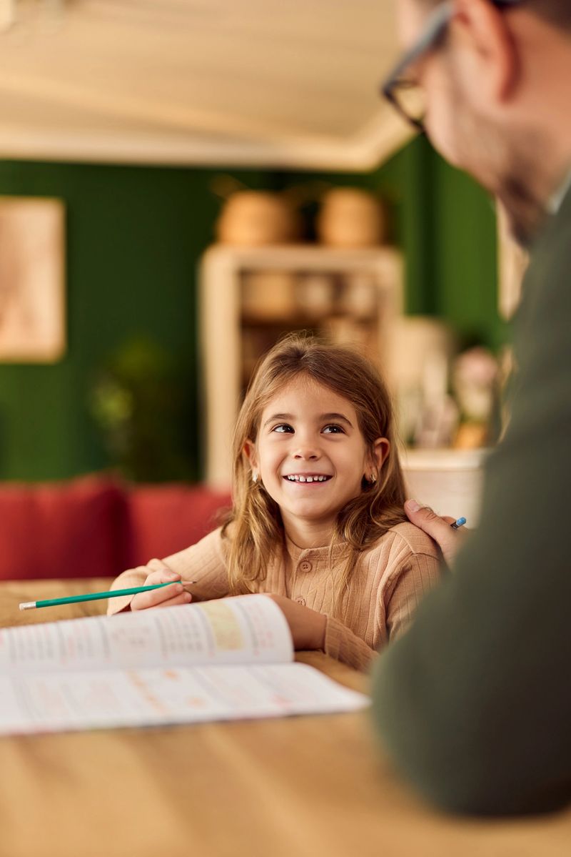 A young girl smiles while studying with her father at a kitchen table.