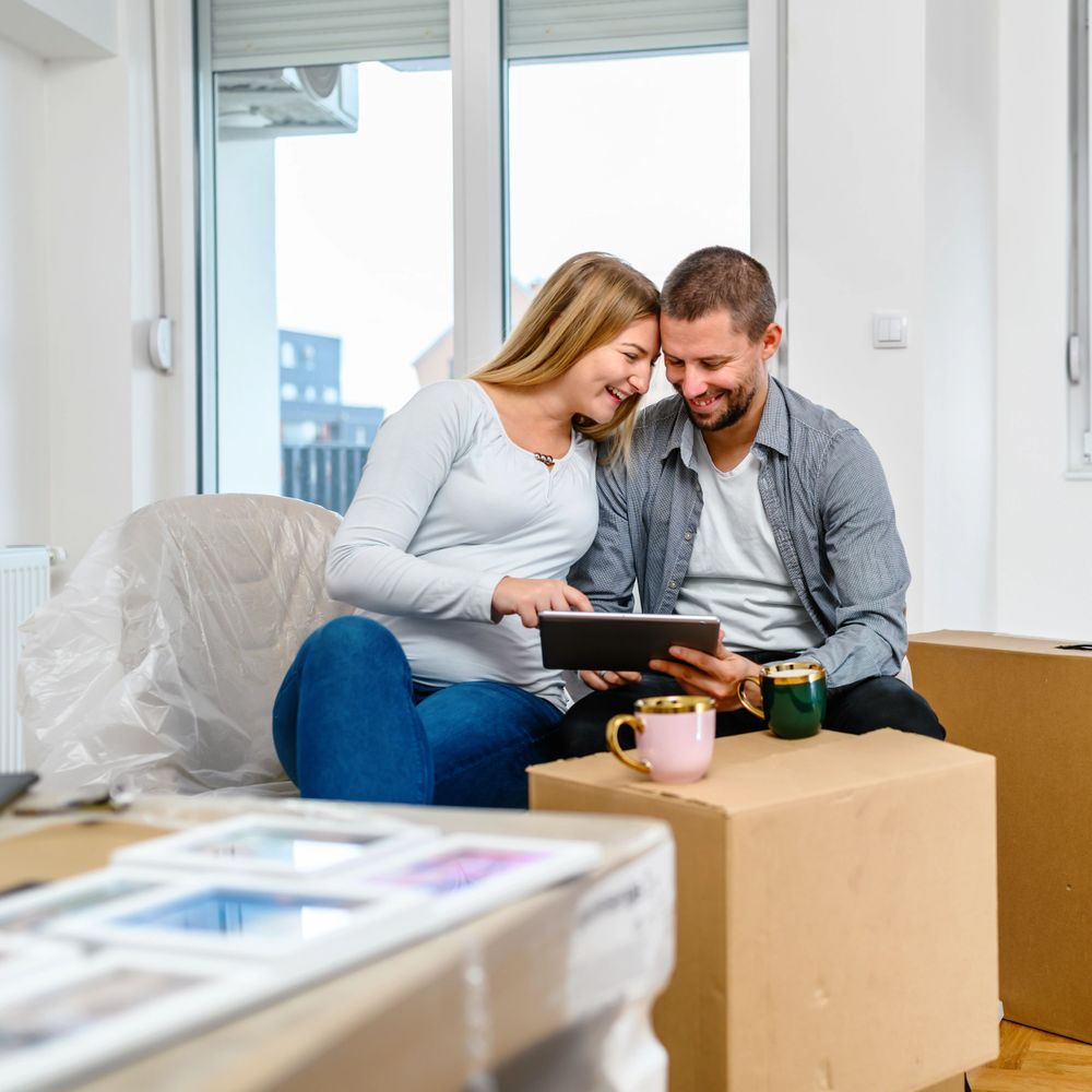 Happy couple sitting on cardboard boxes, looking at a tablet in their new home.