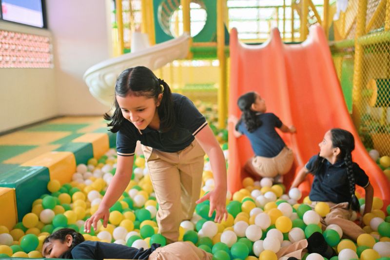 Schoolchildren enjoy a colorful indoor ball pit and slide area, laughing and playing together. Their joyful expressions reflect the importance of play and friendship during school time. Childhood happiness and playful energy in a safe and engaging environment.