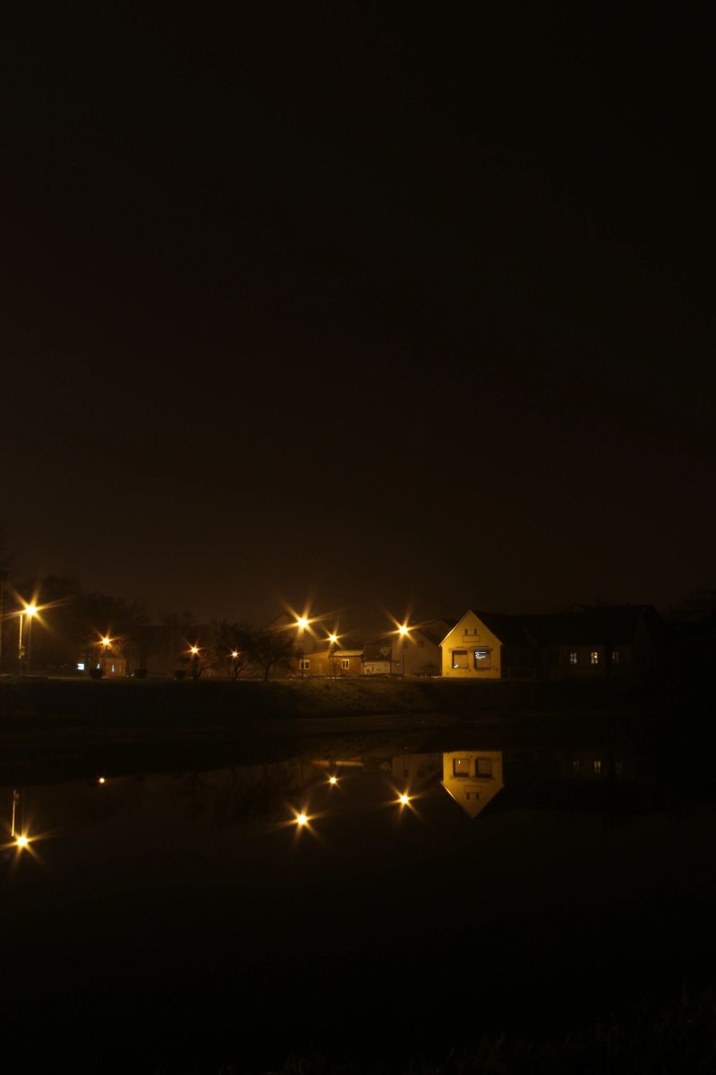 A quiet night scene of a small town street lined with glowing street lamps, their reflections shimmering on the calm water, creating a serene, cozy neighborhood mood.