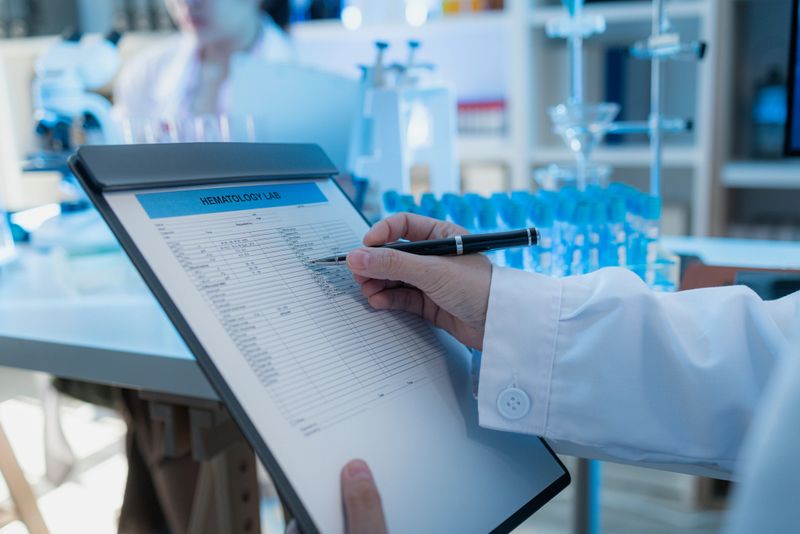 A close-up view of a scientist reviewing hematology lab data on a clipboard in a modern laboratory environment. The desk is filled with glassware, test tubes, and digital devices.