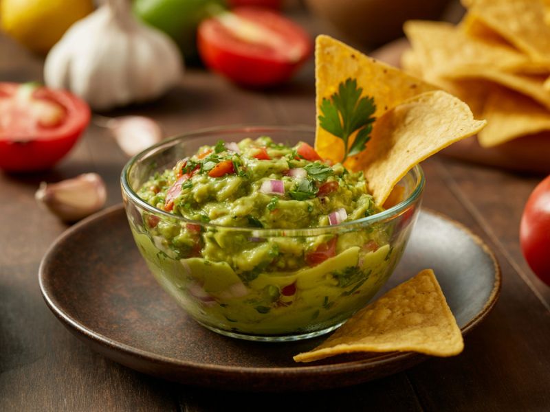 A bowl of fresh guacamole garnished with tortilla chips and surrounded by ingredients like tomatoes and garlic on a wooden table.