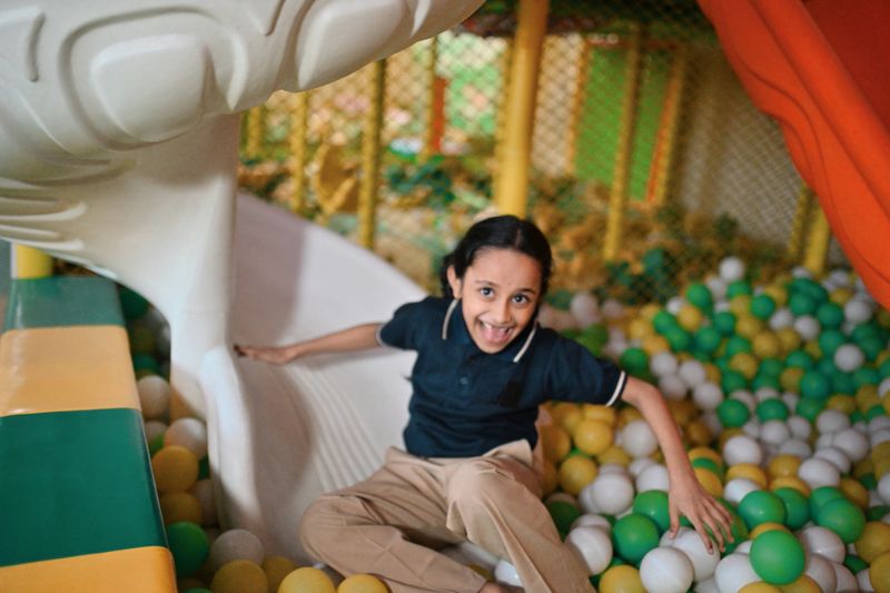 A cheerful primary school girl enjoys sliding into a vibrant indoor ball pit filled with yellow, green, and white plastic balls. Captured mid-laughter, she exudes joy and excitement as part of a fun playtime session. Carefree childhood moments, active play, and school-life balance. Early education, kids’ recreation, happiness, and playful learning environments.