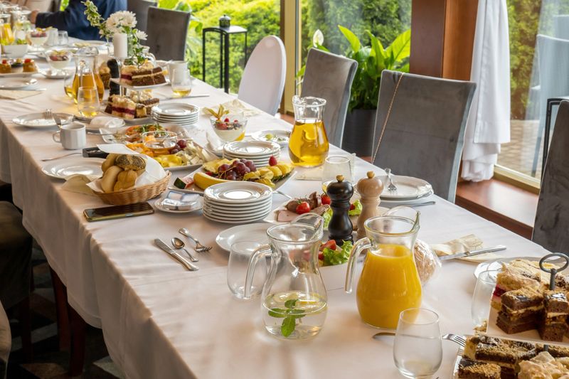 Long banquet table set for hotel event catering with plates, cutlery glassware jugs of juice and water, fresh bread, salads, cold cuts, fruit platters cakes, indoor daylight restaurant reception setup