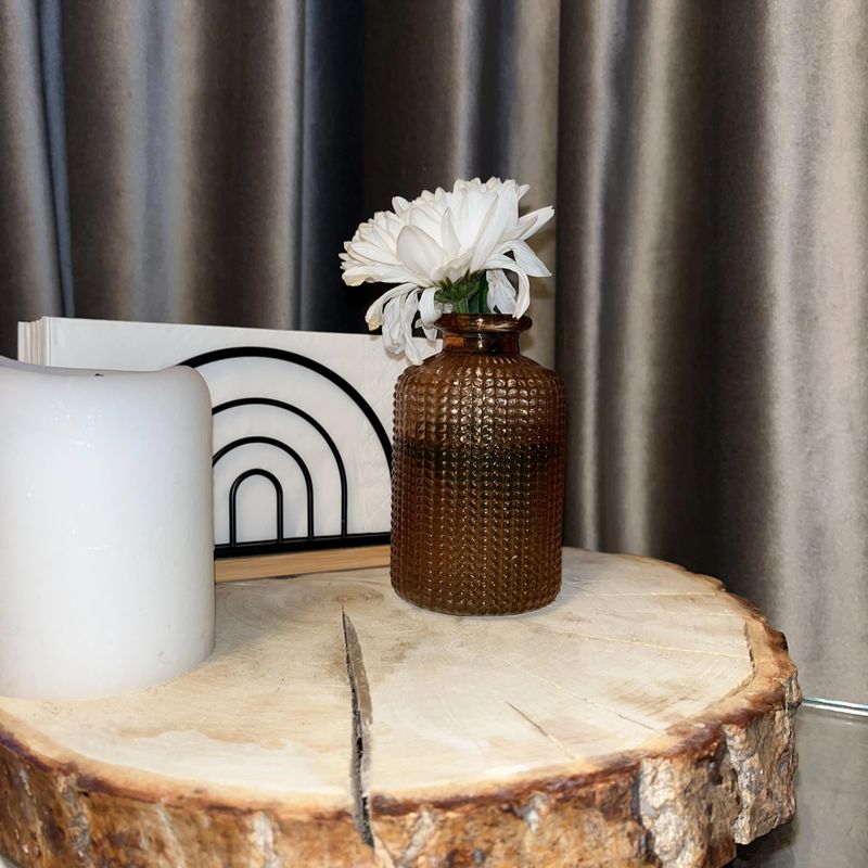 A textured glass vase holds fresh white flowers on a rustic wooden table. Next to it, a white candle and a modern art piece create a cozy atmosphere in the room.