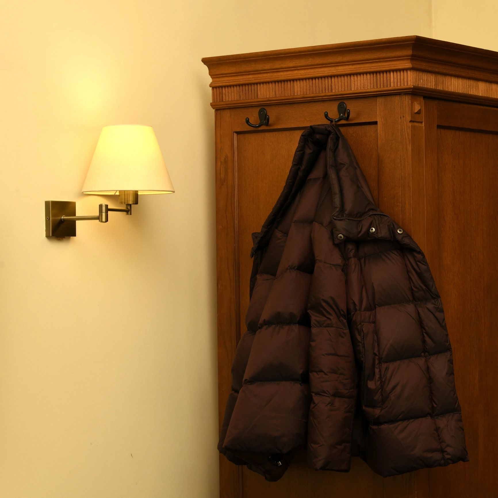 Brown puffer jacket hanging on wooden coat hooks next to a wall lamp.
