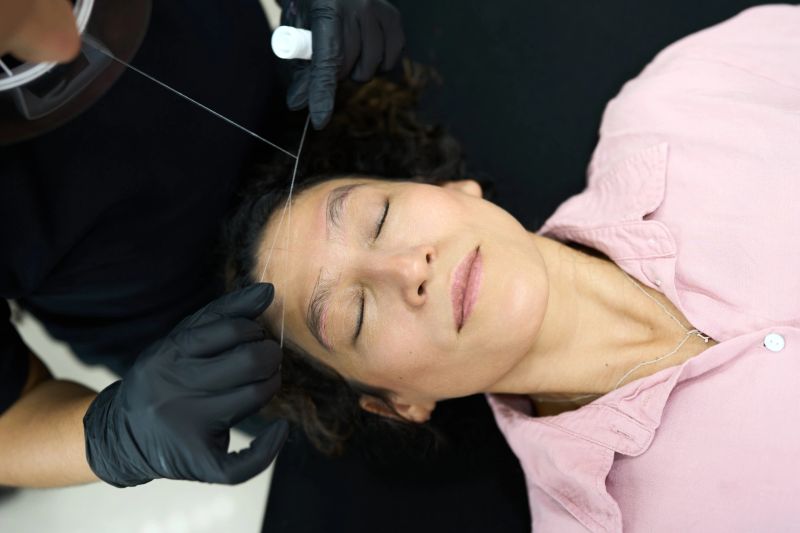Young adult woman receiving professional eyebrow threading service at a beauty salon. Esthetician using thread for hair removal