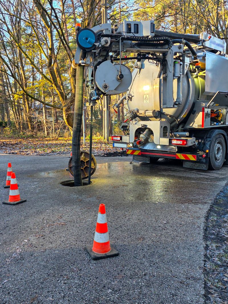 Vacuum sewer maintenance truck cleaning an underground pipeline with large suction hose and safety cones placed around an open manhole on forest road