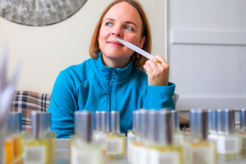 Portrait of a female entrepreneur perfumer smelling perfume at home using a scent strip. In the foreground is an array of perfume bottles arranged on a tray. The woman is sitting on her sofa in the front room. She has piercing blue eyes and wears a blue fleece.