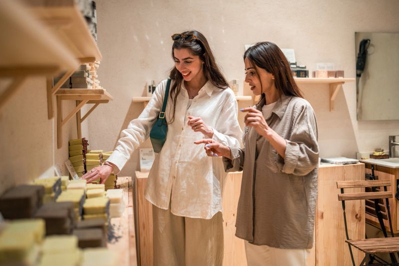 Two diverse female tourists in casual attire browse traditional handmade soaps in a well-lit shop in Al Balad, enjoying the vibrant indoor shopping experience.