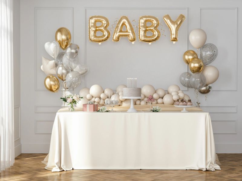 A beautifully decorated baby shower setup featuring gold and silver balloons, a cake on a white stand, wrapped gifts, floral arrangements, and a soft beige dessert table. The elegant display creates a warm and festive atmosphere for celebrating the arrival of a new baby.