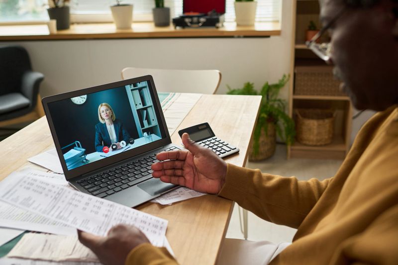 Middle aged Black man holding tax documents consulting young adult Caucasian woman via video call on laptop discussing financial paperwork with calculator on table