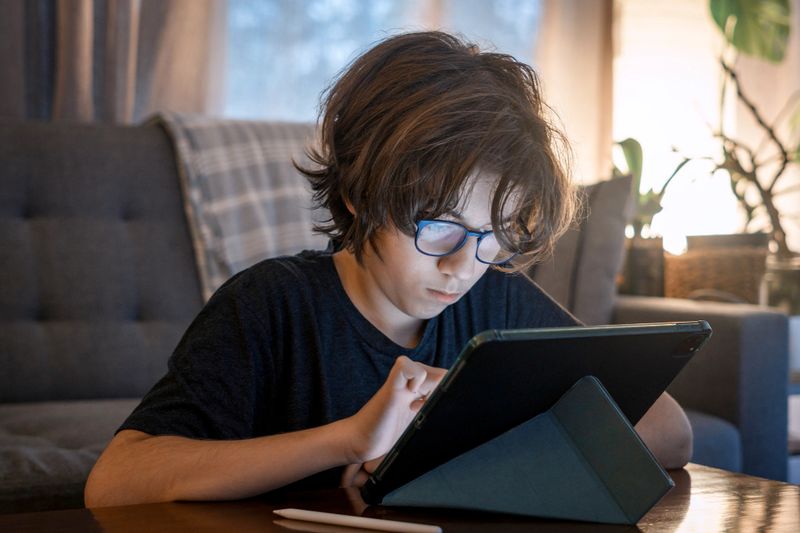 Young boy studying with a digital tablet and stylus, illuminated by blue screen light
