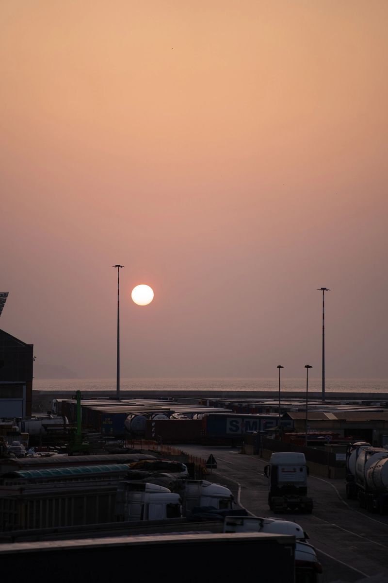 Sunrise over busy port with trucks and tall lampposts in vertical frame, intimate orange sky and soft haze highlighting stacked