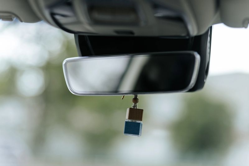 Close-up of a carâs rearview mirror with a small wooden air freshener hanging from it, against a blurred background.