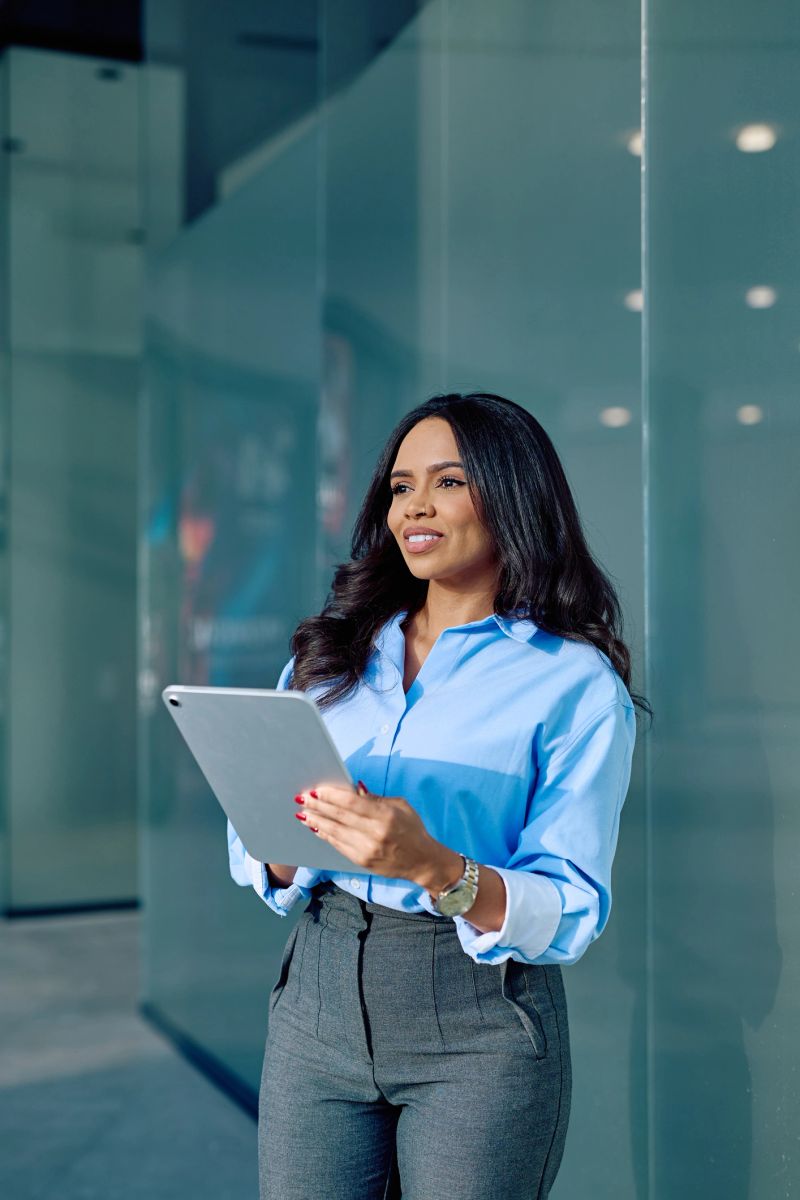 Young adult businesswoman holds a digital tablet, looking away with a thoughtful and positive expression in an urban corporate environment