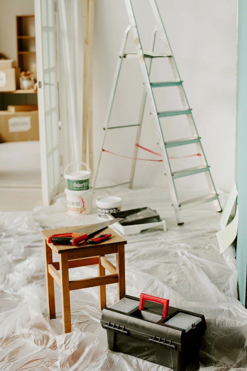 Renovating new home, painting walls and preparing space for moving, plastic sheets covering floor, step ladder, paint cans, toolbox and tools visible in foreground