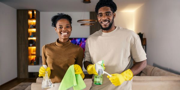 Smiling couple in yellow gloves holding cleaning supplies in a modern living room.