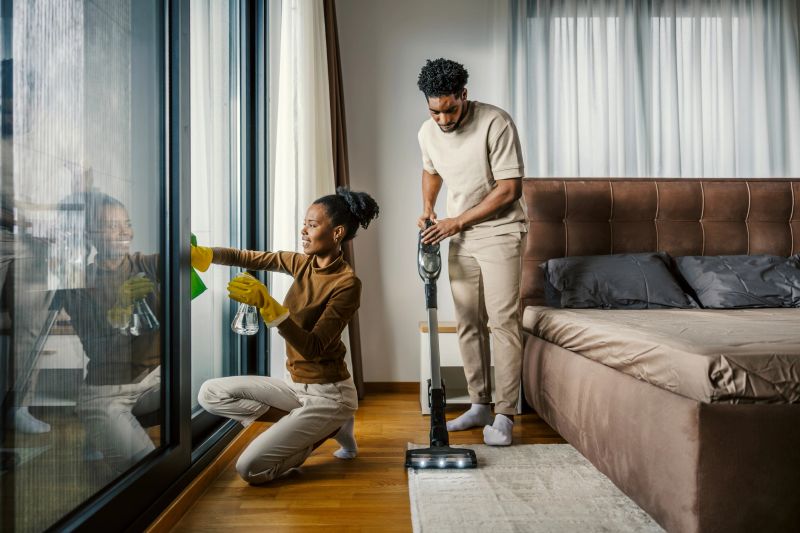Young adult couple collaborating on household chores, a woman washing windows while a man vacuums the bedroom carpet