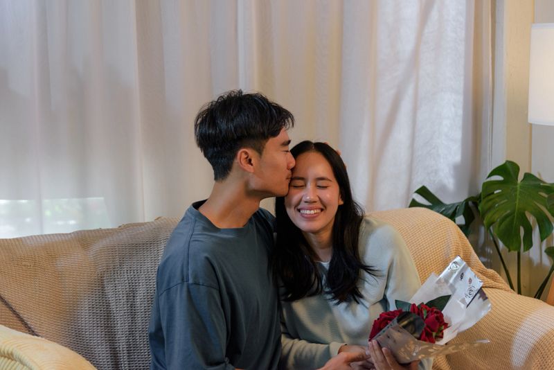 Happy young couple, man woman love home, romantic valentine day, sharing tender moments, soft kiss forehead, receiving beautiful red flowers, celebrating intimate occasion joyfully.