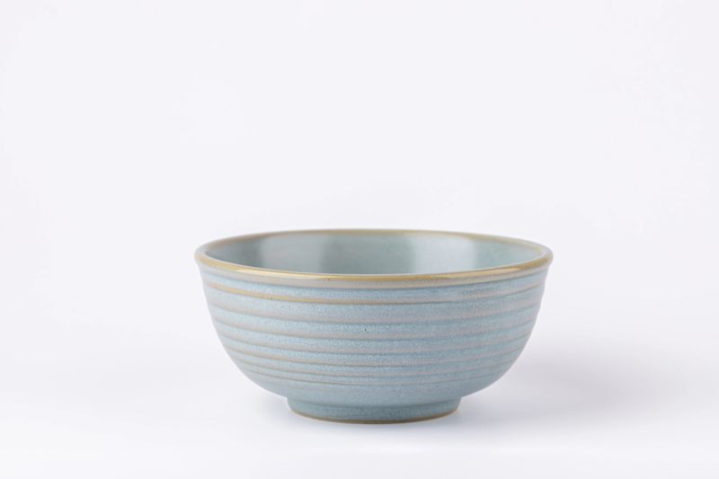 Front view of light blue ceramic bowl with horizontal lines on white background, natural and serene style with copy space. Concept of handmade kitchenware and calm aesthetics