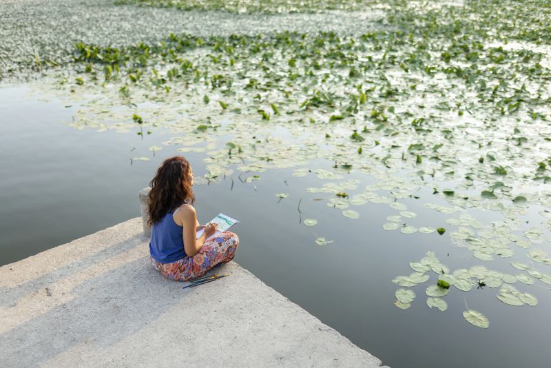Woman is sitting by the lake, enjoying beautiful morning and painting with water colors. She is surrounded with water lily witch is her main inspiration.