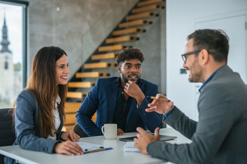 Diverse couple consulting with a professional male adviser in a bright, contemporary office setting, discussing business strategies, finance, or investment plans with engaged expressions