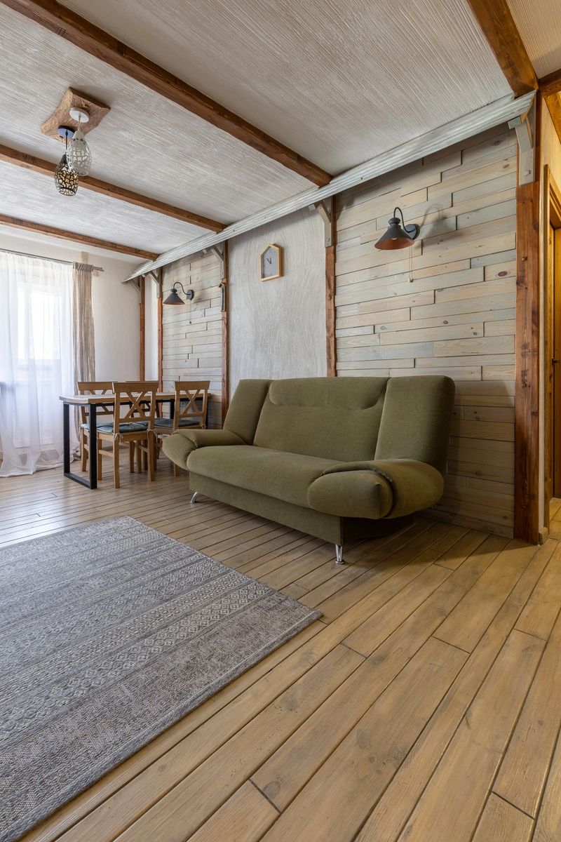 Bright rustic living room with green sofa, wooden walls, dining area, and soft natural light creating a cozy home atmosphere.