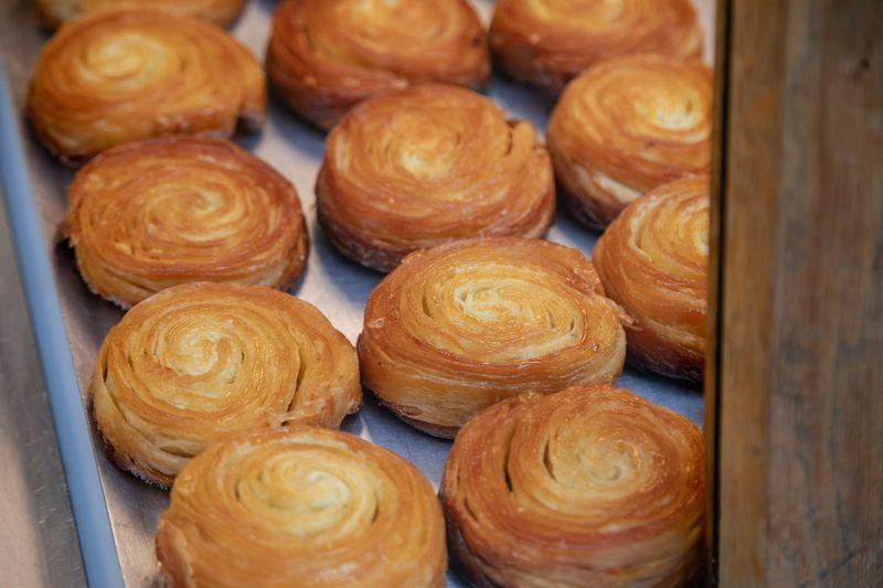 A close-up view of traditional Breton kouign-amann pastries arranged on a metal tray, showcasing their golden, caramelized, buttery layers and crisp, spiral texture in a French bakery setting