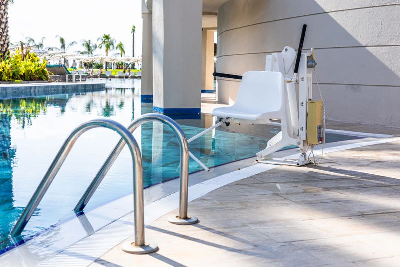 A poolside lift chair for disability people is positioned next to a swimming pool. The area is well-lit with clear blue water and a modern architectural design.