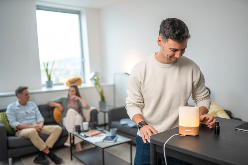 Mid adult Asian man smiling as he adjusts an essential oil diffuser on a sleek table in a contemporary office lounge with colleagues relaxing in the background.