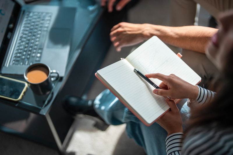 Young Hispanic woman attentively checking notes in a notebook during a business meeting, with a laptop and coffee cup on the table, highlighting her focus and engagement in the discussion.