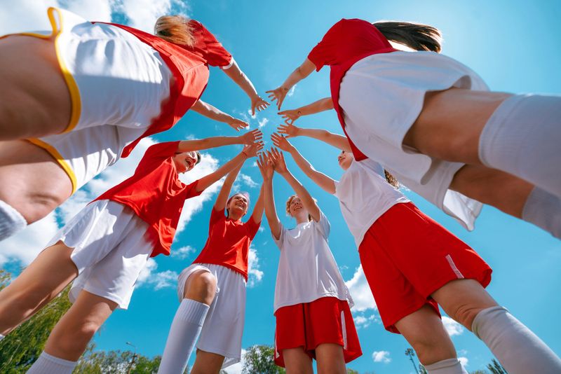 Girls football team in red and white forming team huddle with hands raised under blue sky. Concept of sports unity, youth motivation, girl empowerment, summer team building.