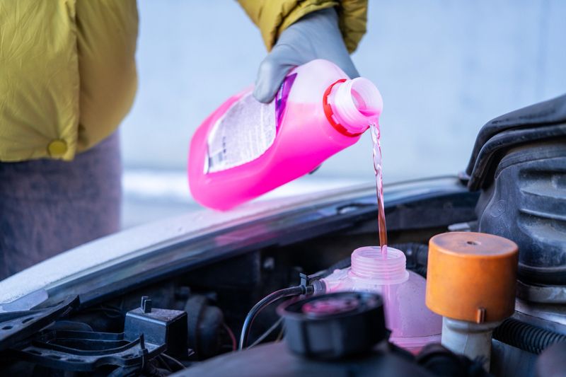 pouring pink coolant into engine reservoir, driver wearing yellow jacket topping up fluid at open car hood close-up of plastic bottle stream filling