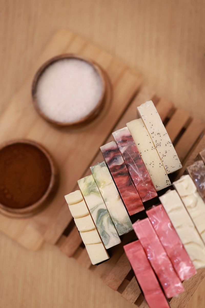 This image highlights a carefully arranged selection of artisanal soap bars placed on a wooden tray within the calming environment of a massage room. The soaps feature varied colors, marbled patterns, and natural ingredients, reflecting the handmade craftsmanship often found in boutique spa settings. Small bowls of salt and powdered botanicals add to the organic and therapeutic atmosphere, emphasizing the use of natural materials in holistic wellness rituals. The warm light, soft textures, and organized layout make this scene ideal for themes related to spa culture, natural skincare, pampering routines, and luxury wellness experiences.