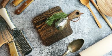 Rustic kitchen utensils arranged around small wooden board with rosemary and grater.