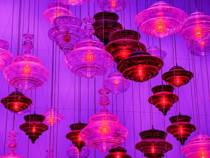 Stunning photograph showcasing dramatic art installation of multiple purple, pink and orange blown glass pendant lights hanging at various heights against blue architectural wall. Features sculptural UFO-shaped and ribbed glass fixtures with warm Edison bulb illumination creating spectacular visual impact. Perfect visual content capturing 2025's biggest trends including statement lighting as art, colorful glass pendants, oversized sculptural fixtures and bold maximalist design. Ideal for interior design inspiration, lighting showrooms, hospitality projects and contemporary architectural content.