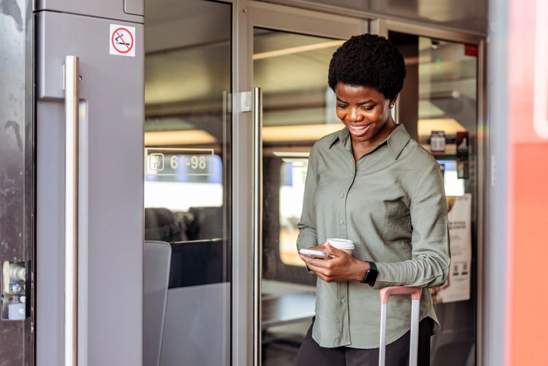 Confident woman holding a smartphone and coffee while stepping out of a stylish train, radiating happiness and positivity, representing travel and technology in a casual and dynamic lifestyle.