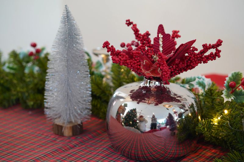 A vibrant and elegant Christmas table arrangement featuring a shiny red and silver vase adorned with berries and holly alongside a mini pine tree on plaid cloth.