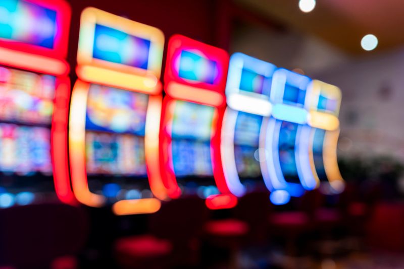 Defocused casino slot machines glow with colorful neon frames, forming a soft bokeh pattern across the gaming floor. The blurred lights create a versatile abstract background, ideal for gambling concepts, nightlife scenes, entertainment marketing, web headers and modern design projects.