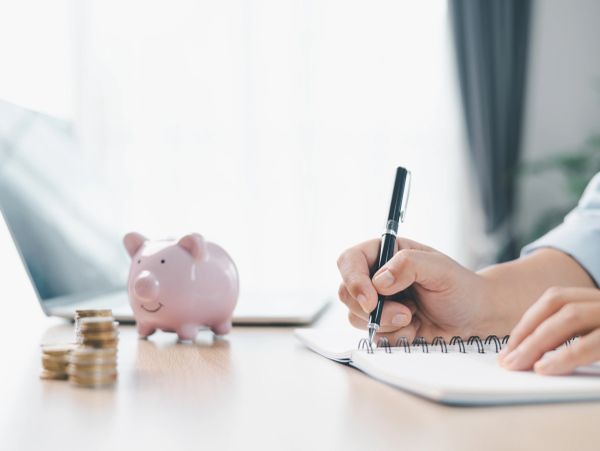 Person writing notes near piggy bank and coin stacks.