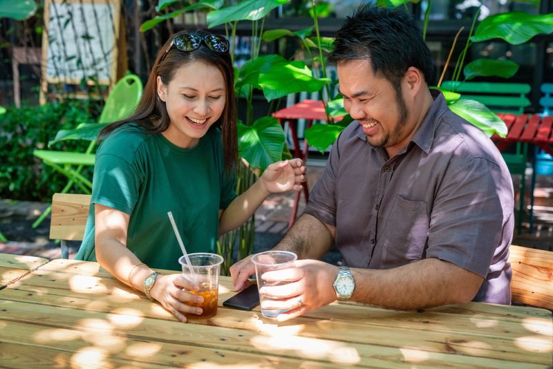 Cheerful couple shares drinks at an outdoor café, laughing and interacting in a bright garden environment. The natural greenery and casual mood create a friendly, warm, and relaxed social lifestyle scene. Kuala Lumpur, Kuala Lumpur Federal Territory, Malaysia, Southeast Asia
