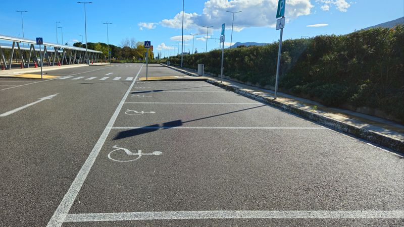 Outdoor parking facility featuring a large canopy structure covered in solar panels, providing shade for vehicles and generating renewable energy. The paved lot has clearly marked lines, including accessible parking spaces, directional arrows, and clear signage under a bright, blue sky.