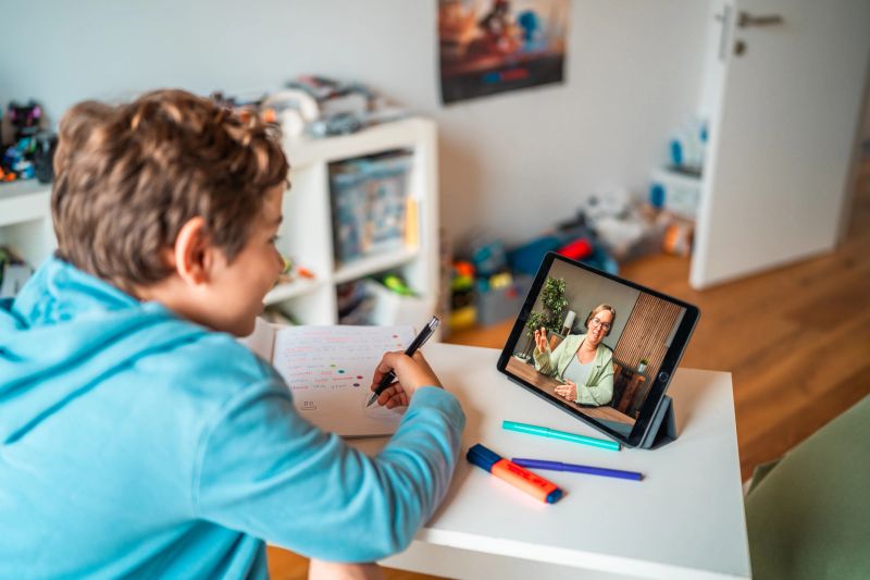 A young Hispanic male concentrating on a virtual learning session at home, interacting with a tutor via digital tablet while taking notes. The study environment includes colorful markers and a well-organized desk, creating a focused and productive setting.