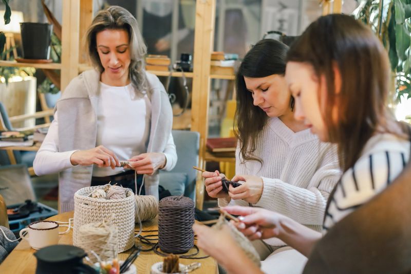 Group of women focused on yarn crafting during a creative workshop, working with hooks and textured fibers in a warm, cozy space that highlights teamwork, hobby and handmade skills.