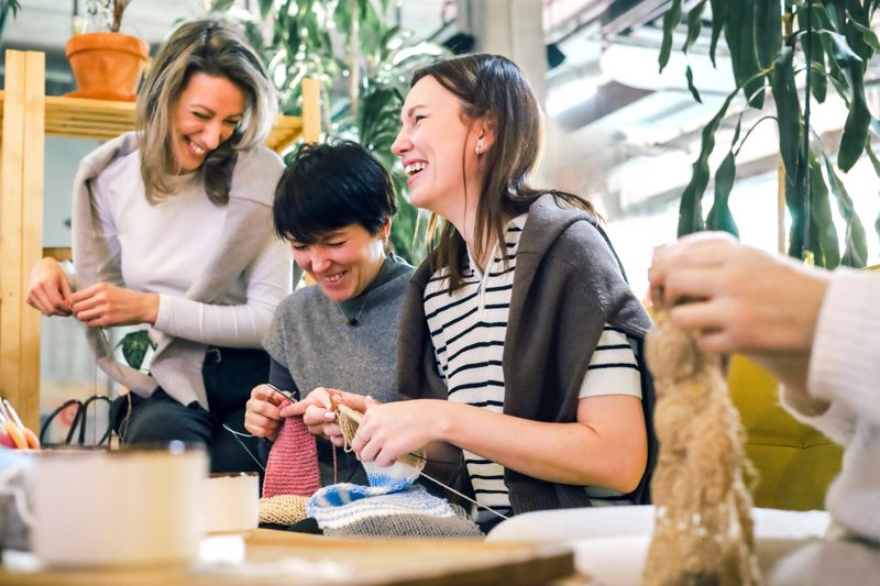 Joyful women knitting together in a cozy creative workshop, laughing, learning, and enjoying a warm handmade atmosphere perfect for lifestyle, craft, hobby, and community-focused commercial use.