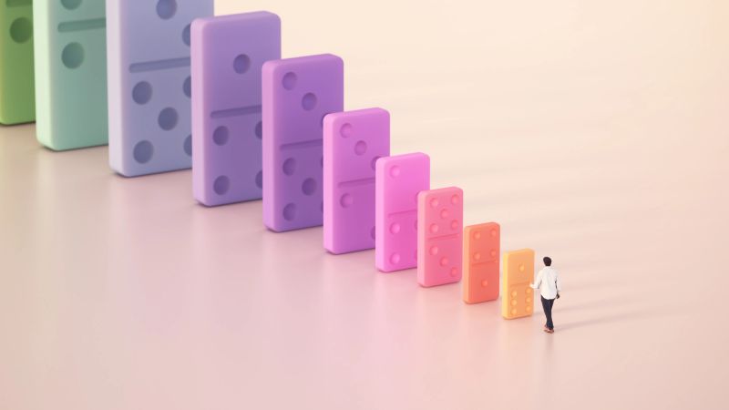 A person strolls toward a series of large, colorful dominos arranged in a descending line. The setting is minimal and bright, emphasizing the vibrant colors of the dominos. Leadership concept.