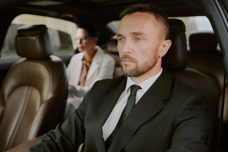 Caucasian middle aged man driving car with serious expression, businesswoman with dark hair and glasses sitting in back seat, both dressed formally, interior of luxury vehicle