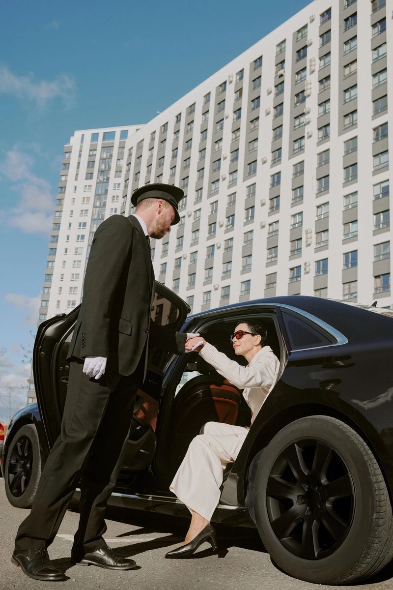 Caucasian middle aged man in chauffeur uniform assisting Caucasian middle aged woman exiting luxury car in urban setting, both engaging in formal interaction outside modern high rise building
