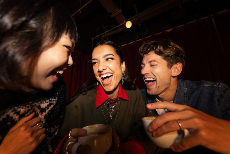 Shot with a flash on camera, this tight image of three people laughing and holding coffee cups is set in a cinema cafe. They are casually dressed in a trendy style. The background is dark red velvet.Videos of this scenario are also available.